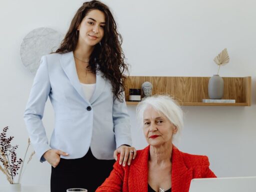 Two businesswomen, one senior, collaborating at a desk in a modern office with laptops and documents.