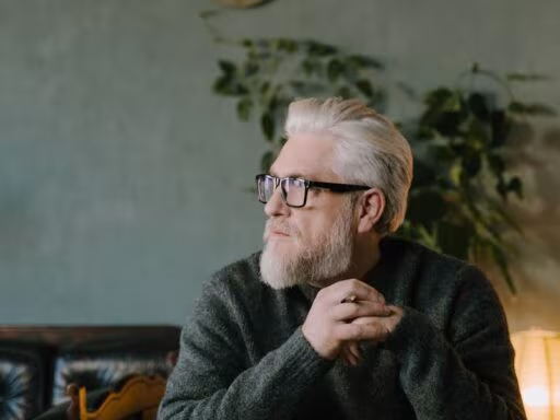 Futur du travail, Elderly man with white hair and glasses sits thoughtfully at a wooden desk in a cozy indoor setting.