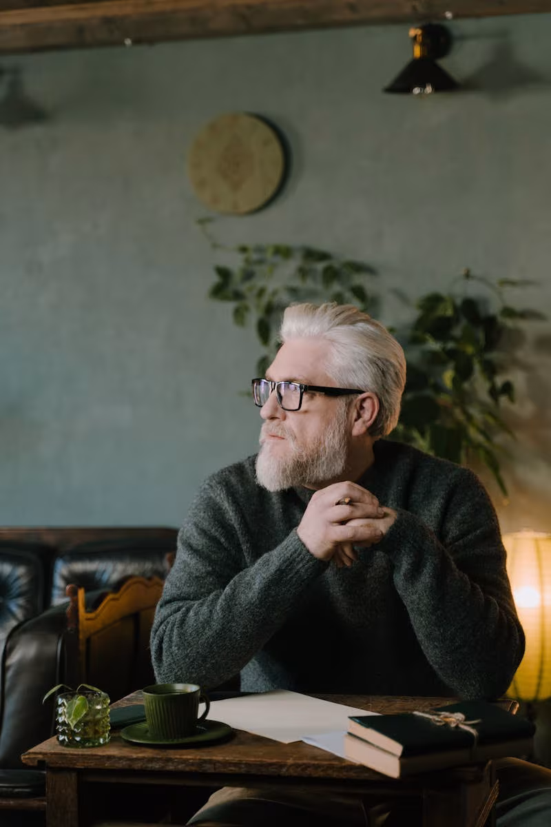 Futur du travail, Elderly man with white hair and glasses sits thoughtfully at a wooden desk in a cozy indoor setting.